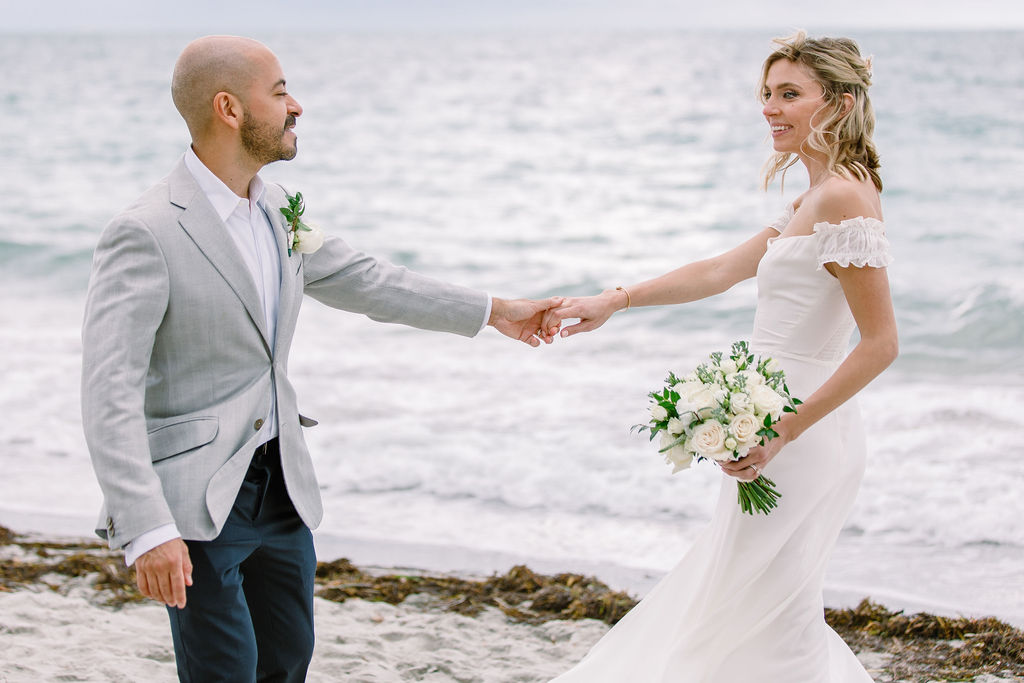 Couple holding hands on the beach with bouquet and ocean waves